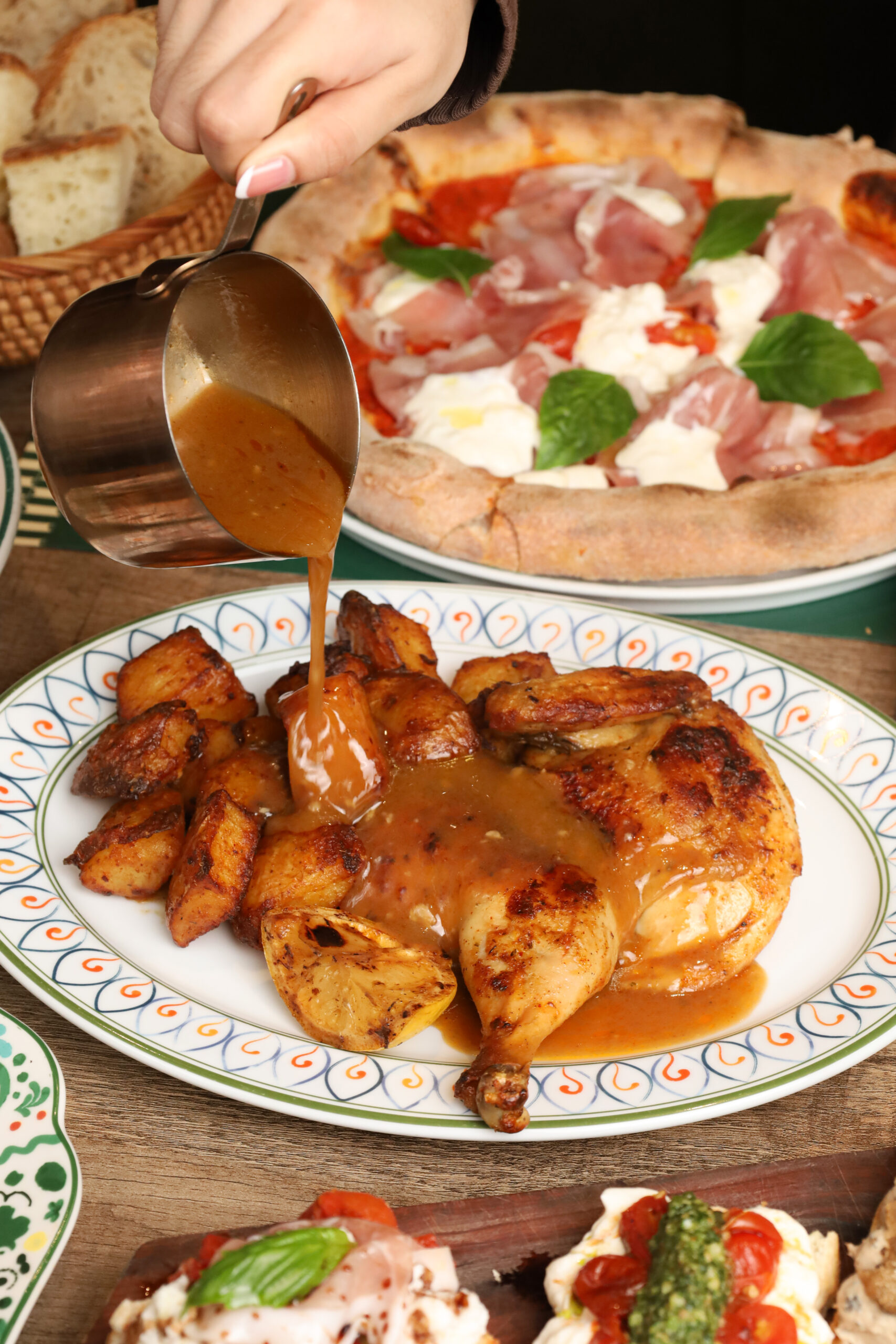 Gravy poured over roast chicken with prosciutto pizza behind, at Bianca Bangkok